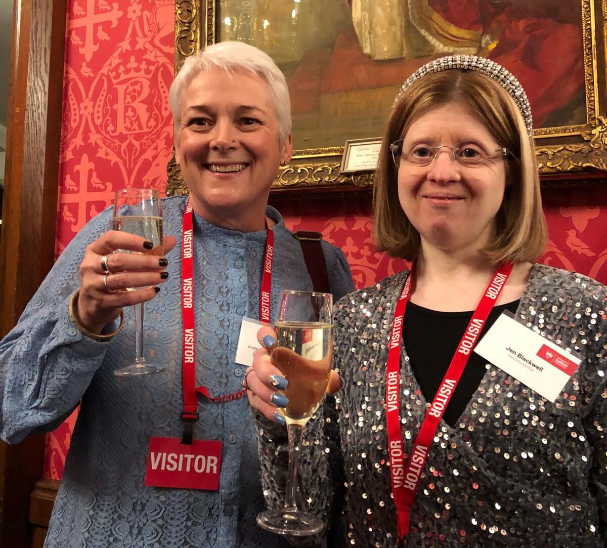 Woman with white hair next to woman with sparkly top and headband raising their glasses and smiling at the camera