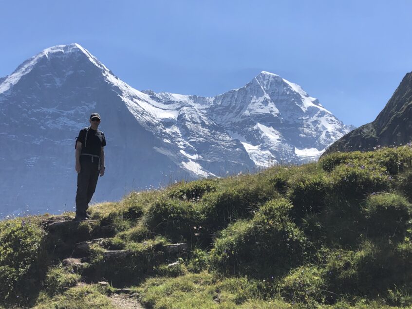 Man stood on grass mountain top with snow covered mountains in background