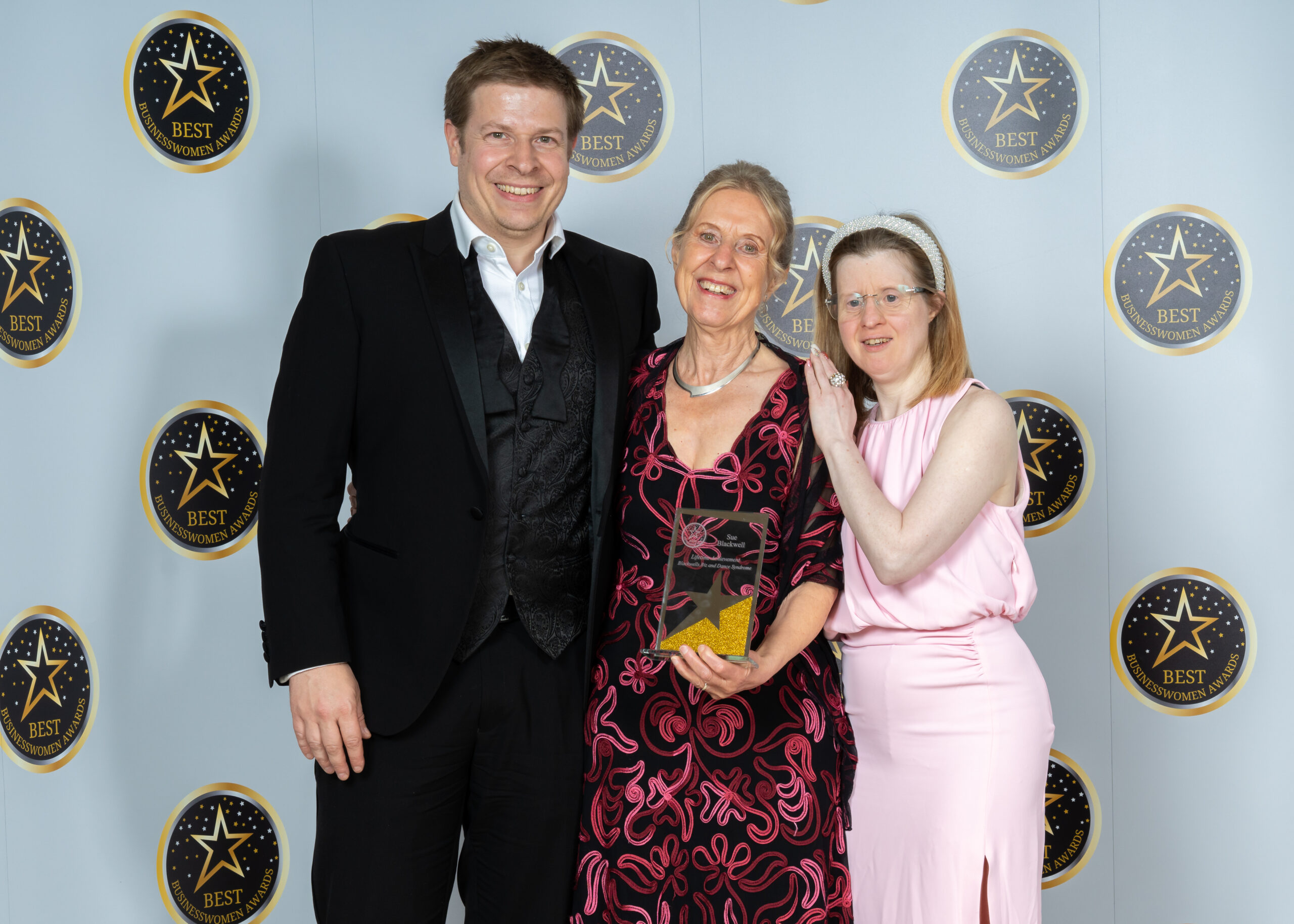 Man in suit with older woman holding award and younger woman resting her hand on shoulder of older woman. All smiling and happy