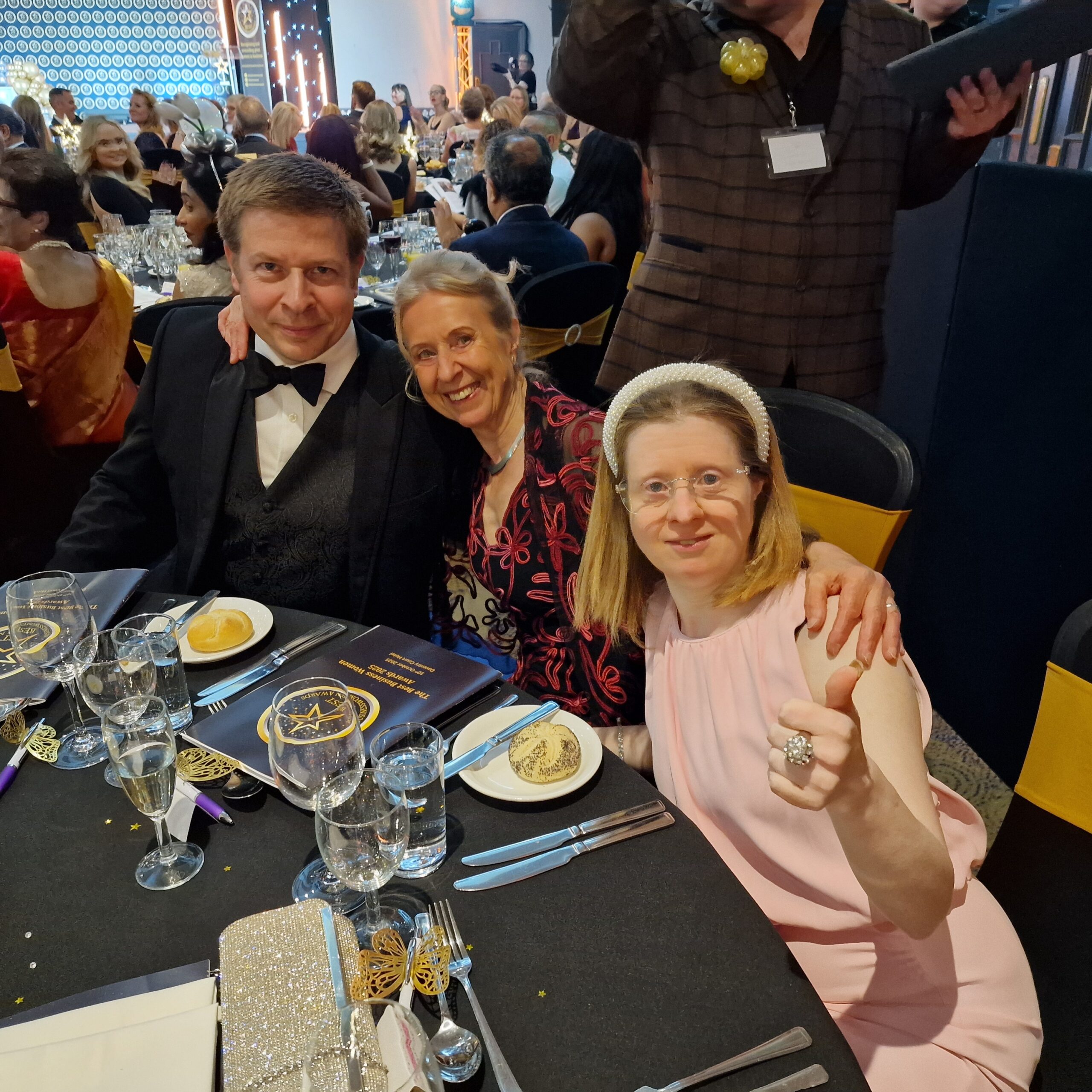 Man wearing black bow tie and suit woman in red dress and younger woman in pink dress and sparkly headband all smiling sat at a dinner table