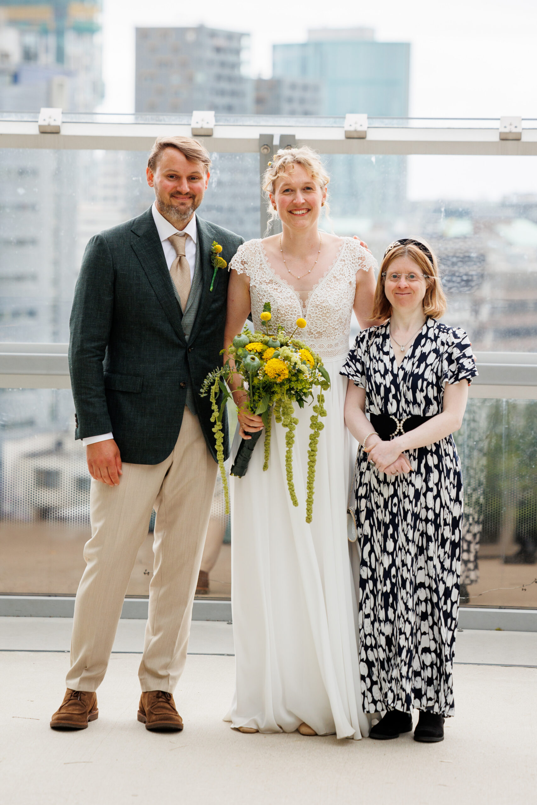 Man in suit and tie, lady in wedding dress holding yellow and green bouquet with shorter lady in black and white dress. All smiling