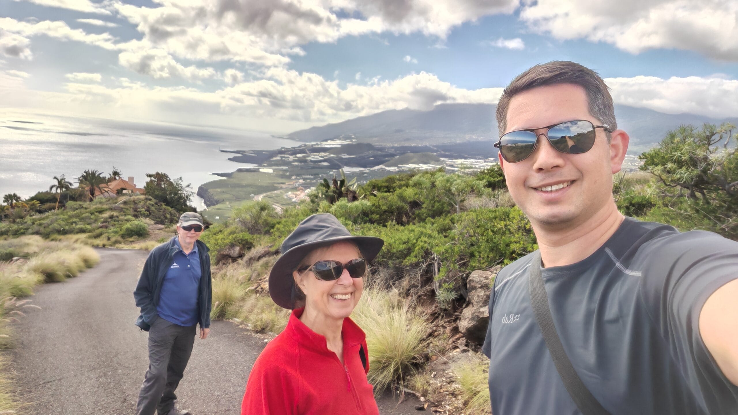 2 men and 1 woman on road wearing sunglasses wtih sea and mountains behind them