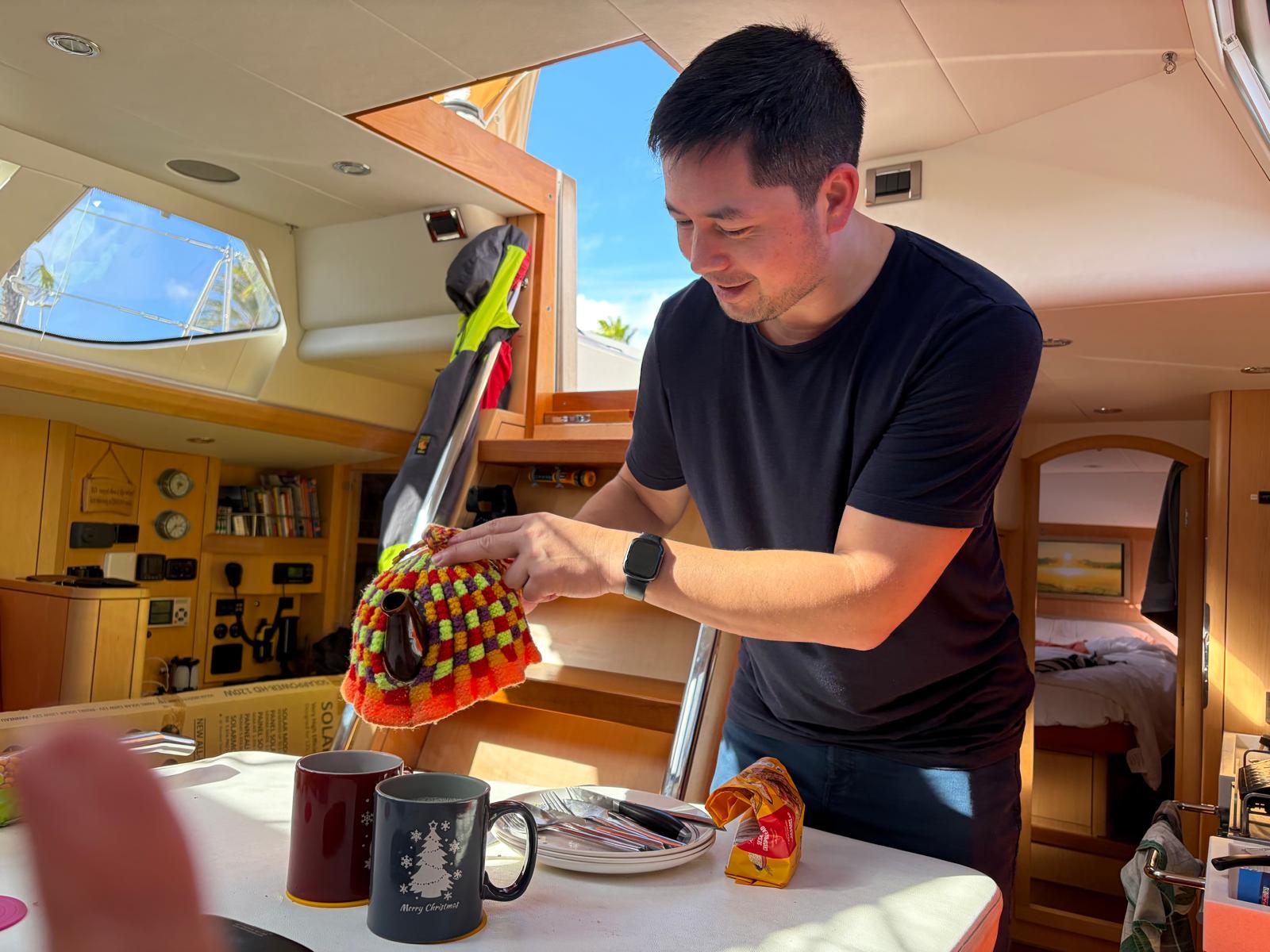 Man with dark hair wearing dark t-shirt pouring tea from a teapot with knitted tea cosy into 2 mugs inside a yacht