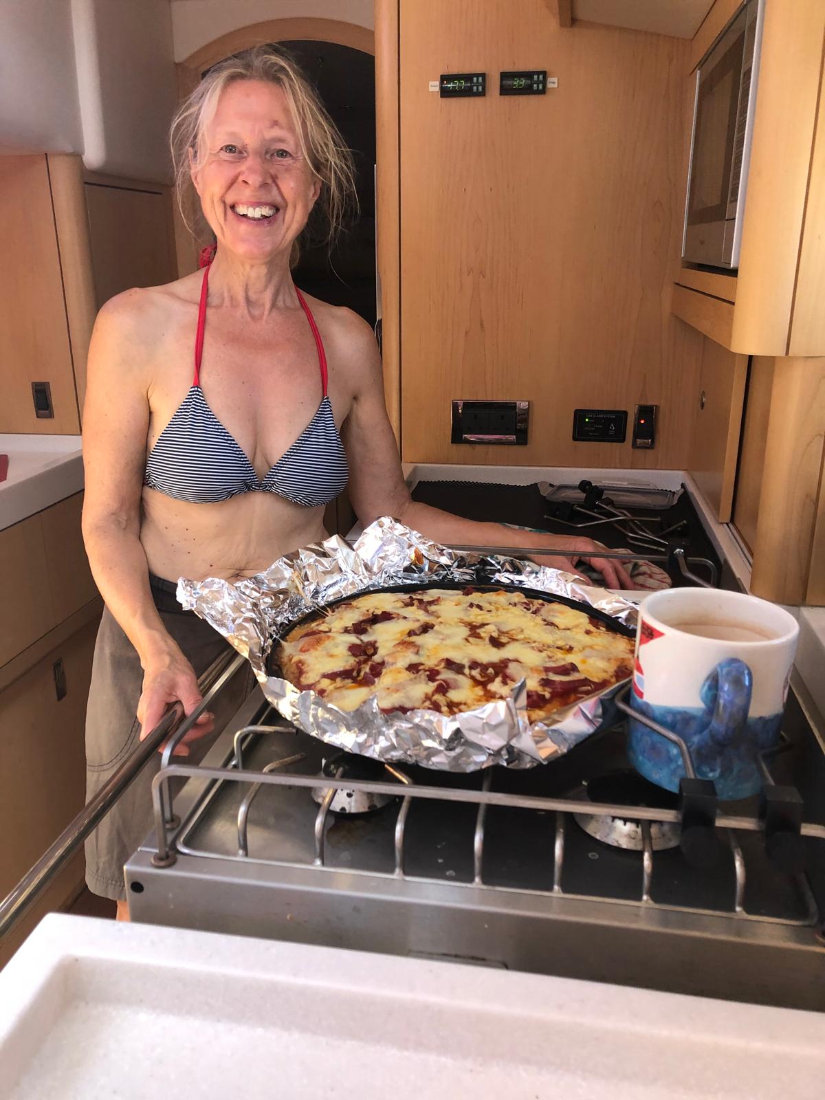 Woman with fair hair wearing bikini top with big pizza on top of an oven on a yacht