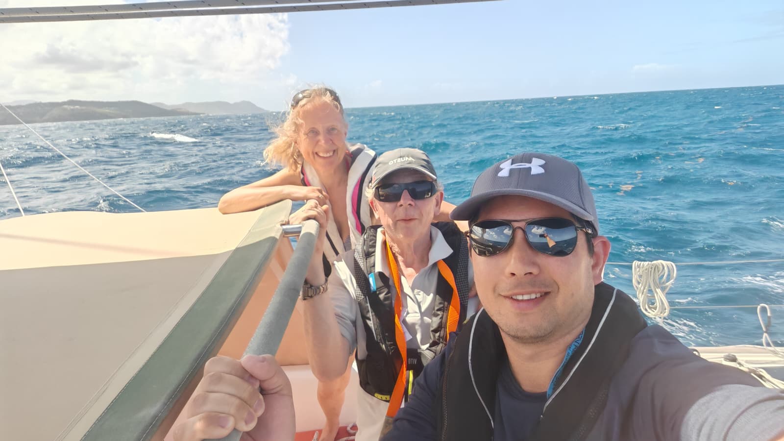 Female with golden hair flying in the wind, with two men wearing caps and sunglasses, all wearing lifejackets on board a yacht with calm sea