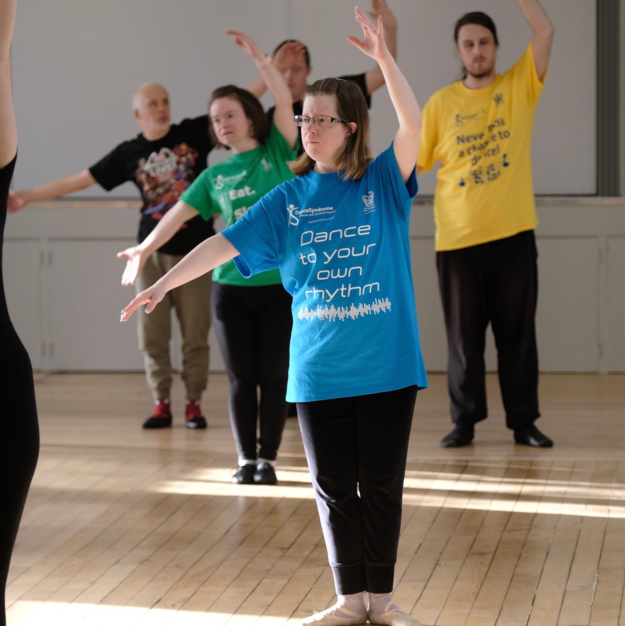Dancers in a room with wooden floor standing straight with one arm out to the side and the other over their heads practicing a dance move