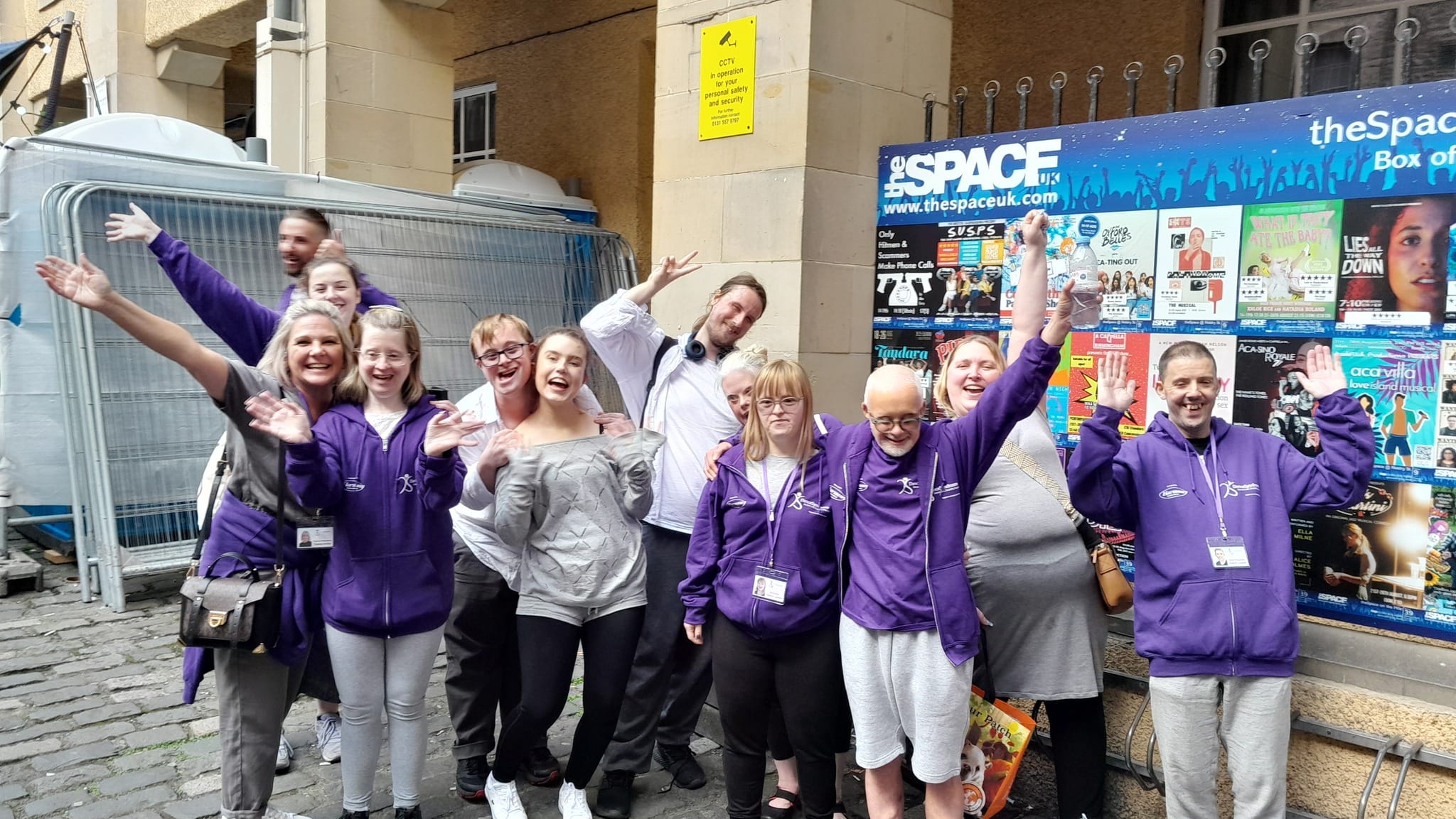 Group of people all smiling with hands up next to noticeboard at Edinburgh Fringe Festival 2023