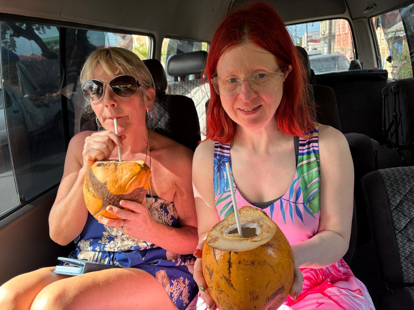Donna with sunglasses on and Jen with glasses on drinking through straws from coconuts in the back of a car