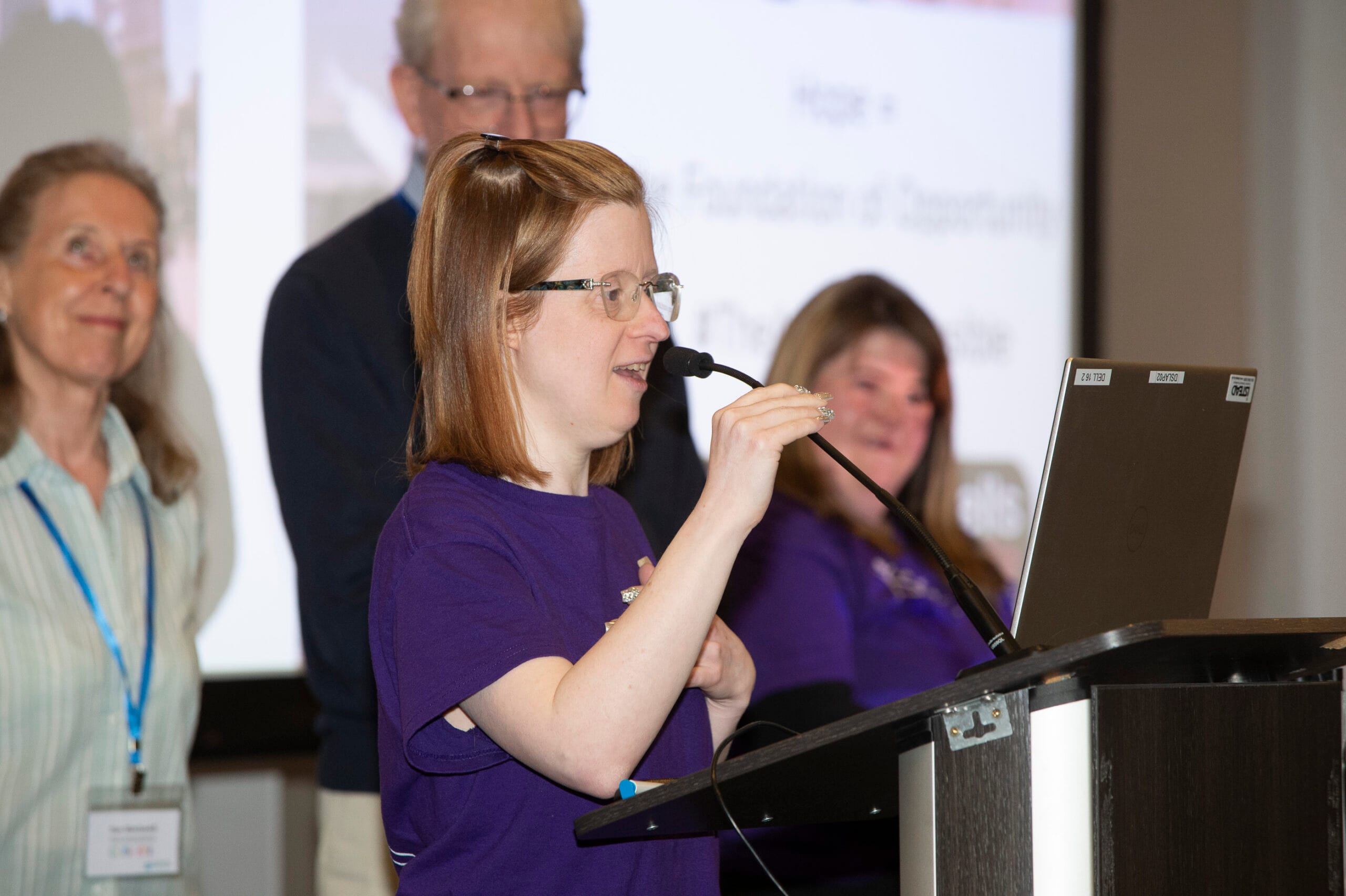 Lady speaking into microphone with 2 ladies and 1 man behind her on stage
