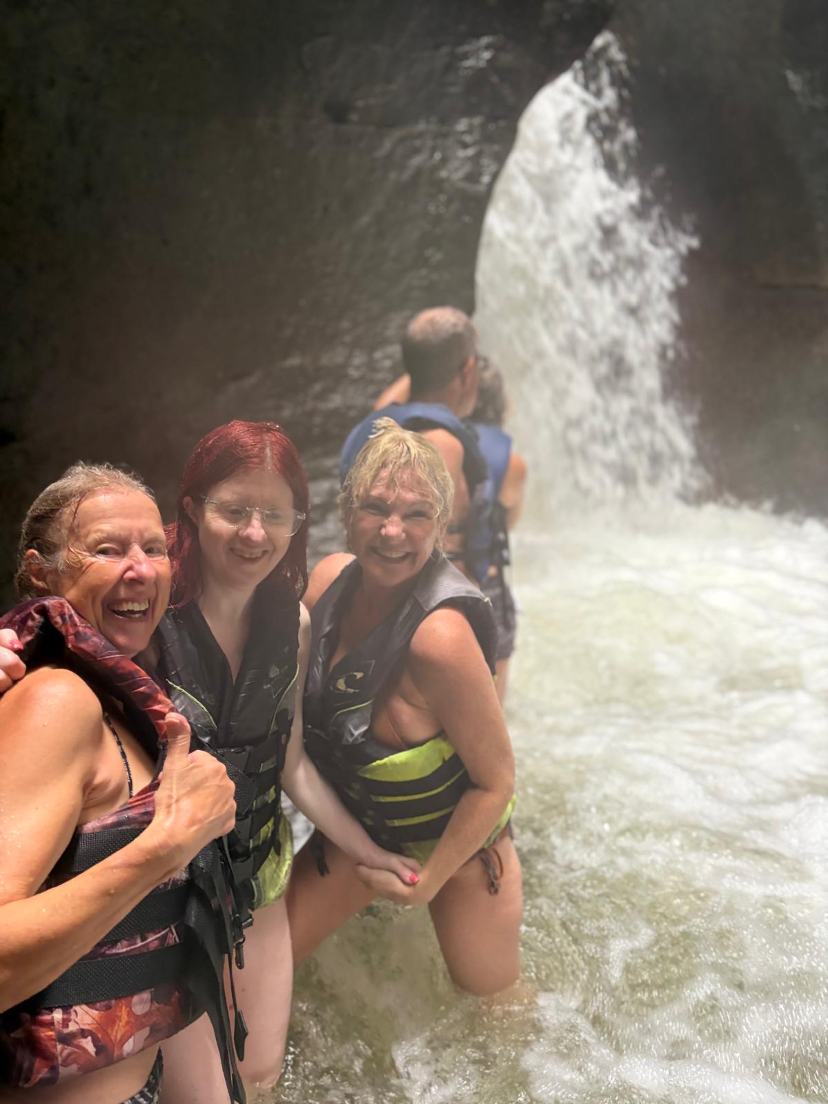 Sue Jen and Donna facing the camera wearing swimwear and lifejackets all with wet hair standing in front of a waterfall.