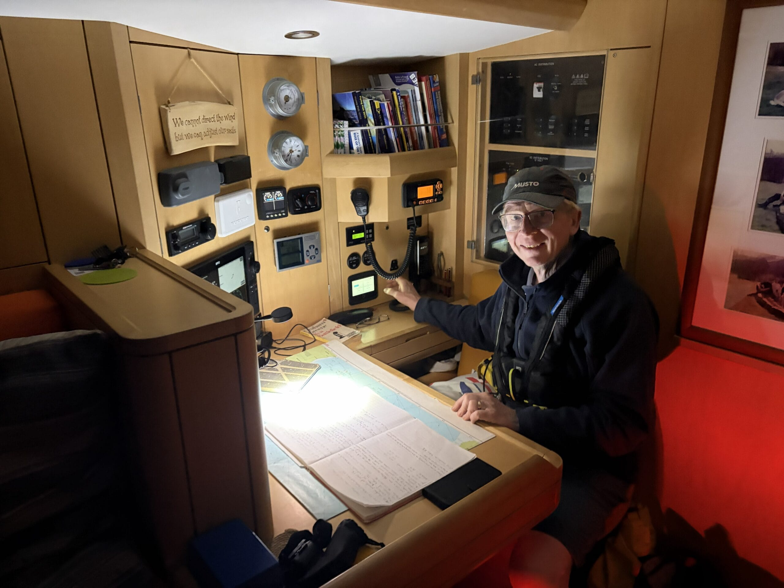 Man with cap sitting in dark with a light on with sailing charts on the chart table in front of him