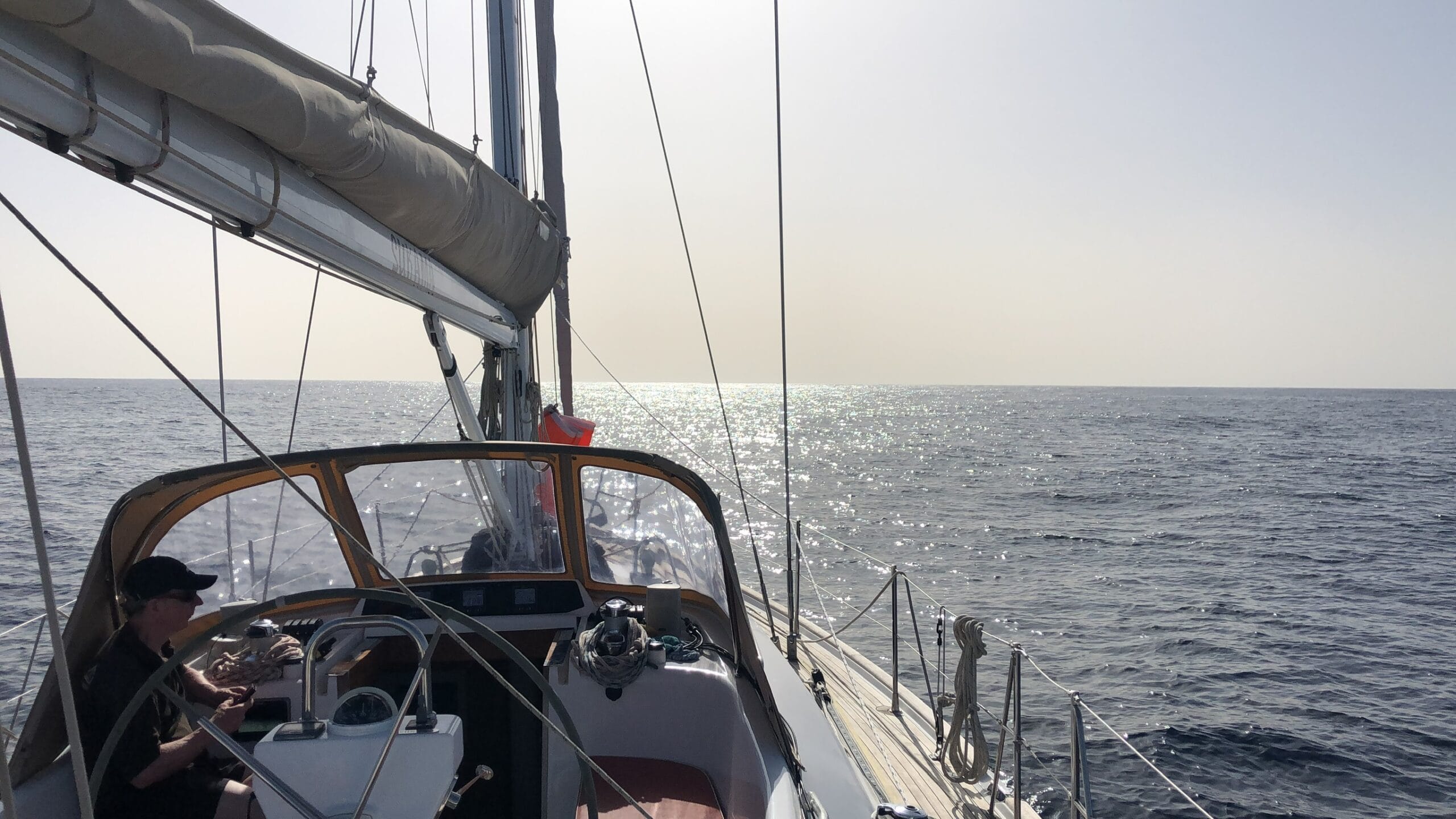 Yacht with sails down motoring on a flat calm sea. Man in cockpit taking shelter from the sun