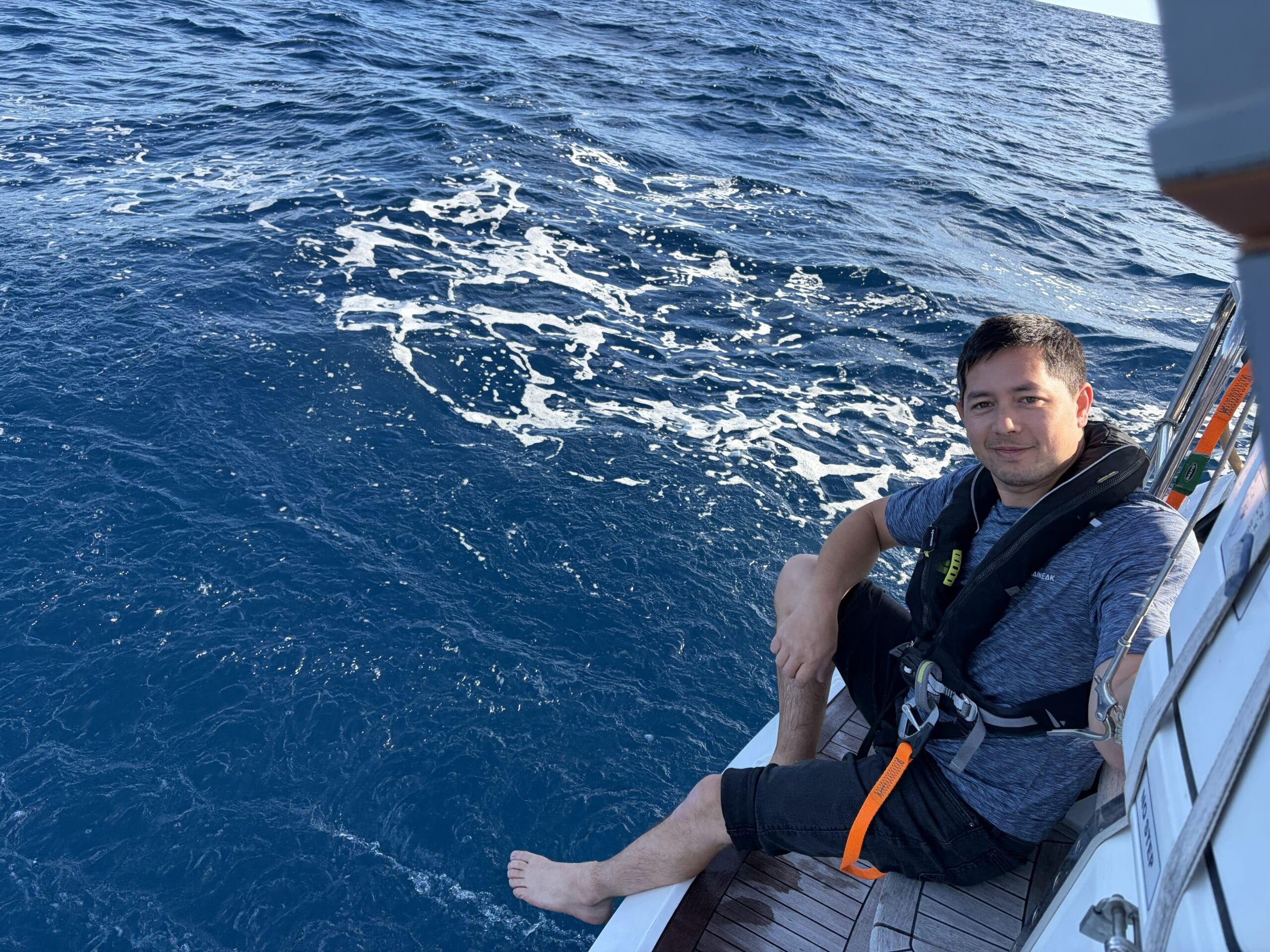Young man with dark short hair wearing life jacket sitting on back of yacht with feet in water on calm sea