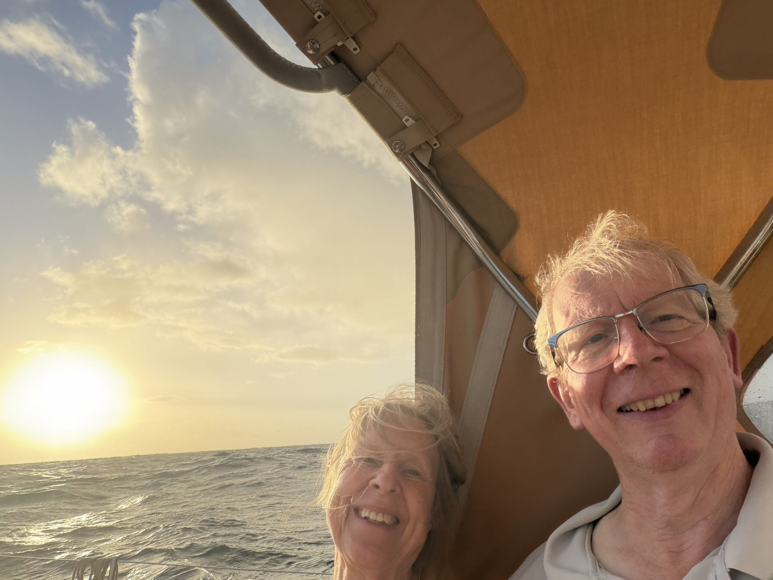 Two people smiling in cockpit of yacht with setting sun falling into the open ocean to the left of them