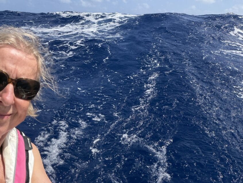 Lady with fair hair and sunglasses at back of boat with a wave high behind her