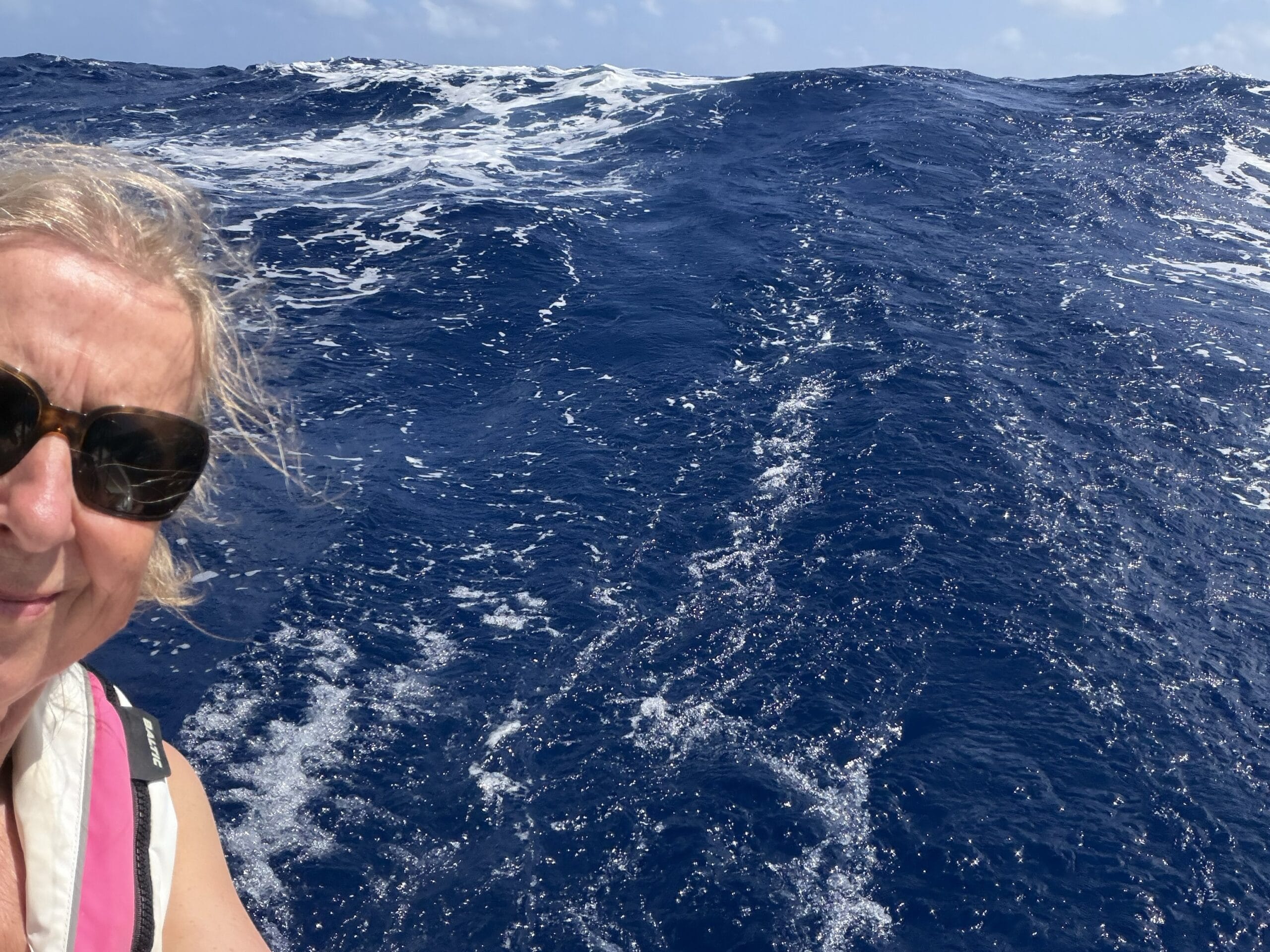 Lady with fair hair and sunglasses at back of boat with a wave high behind her