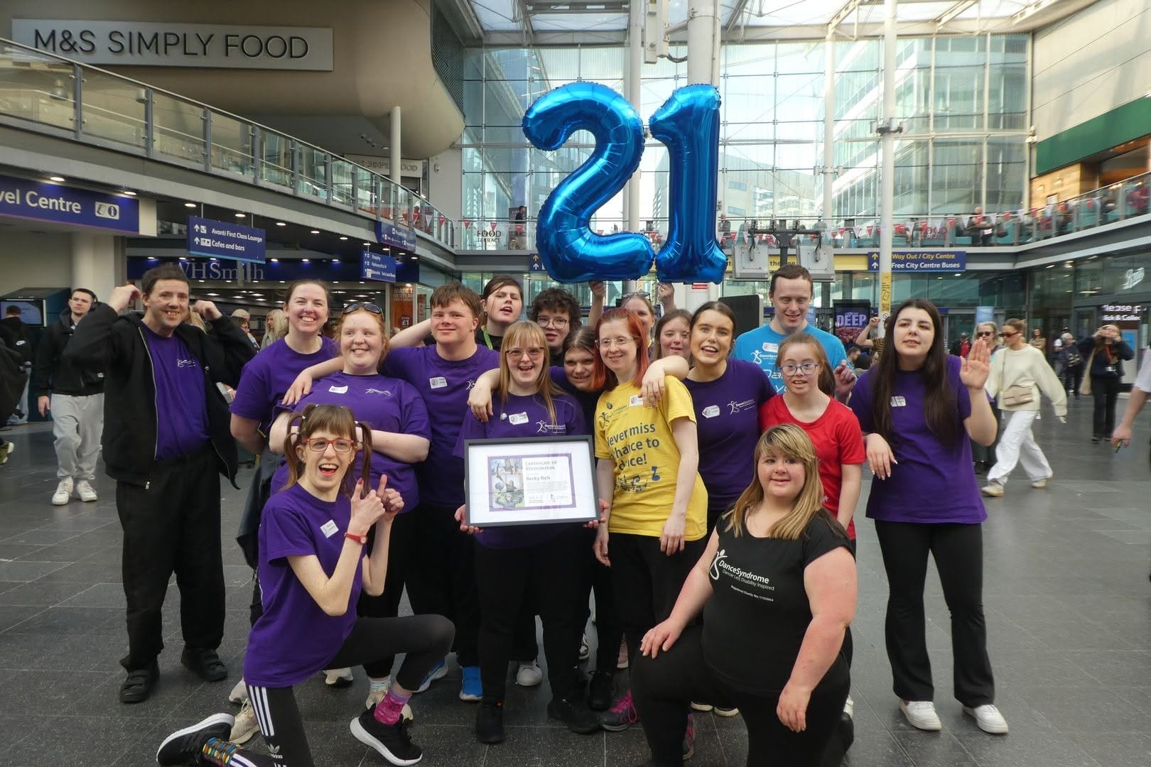 Group of DanceSyndrome dancers at Manchester Piccadilly station with Becky and Jen in the centre. Balloons that read 21 for Trisomy 21 Down's syndrome the day that they were dancing and celebrating 